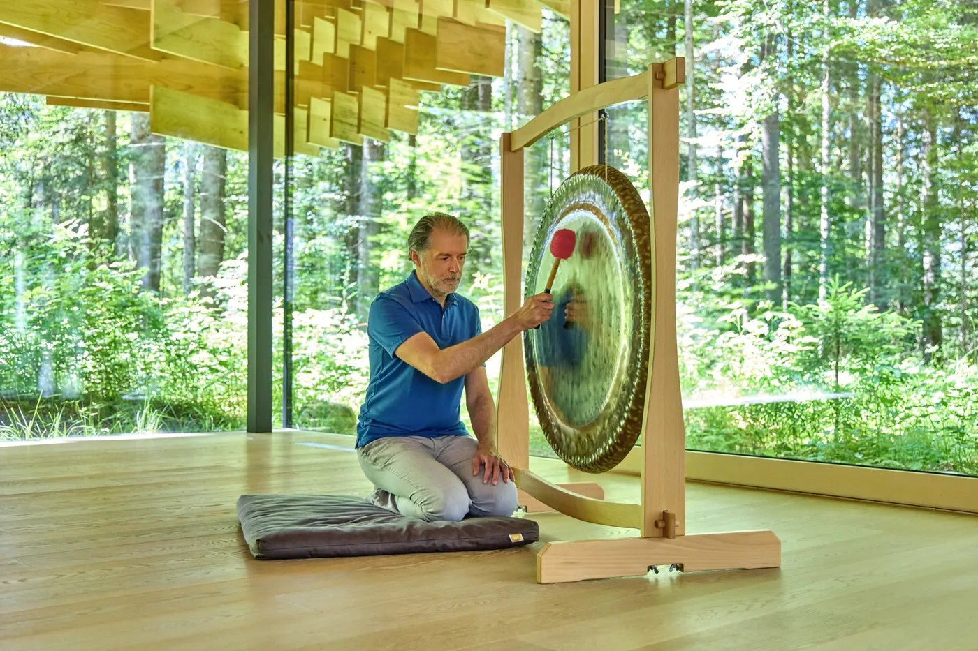 Alexander Renner spielt den Gong in einem hellen Holzraum mit großer Glasfront und Waldblick