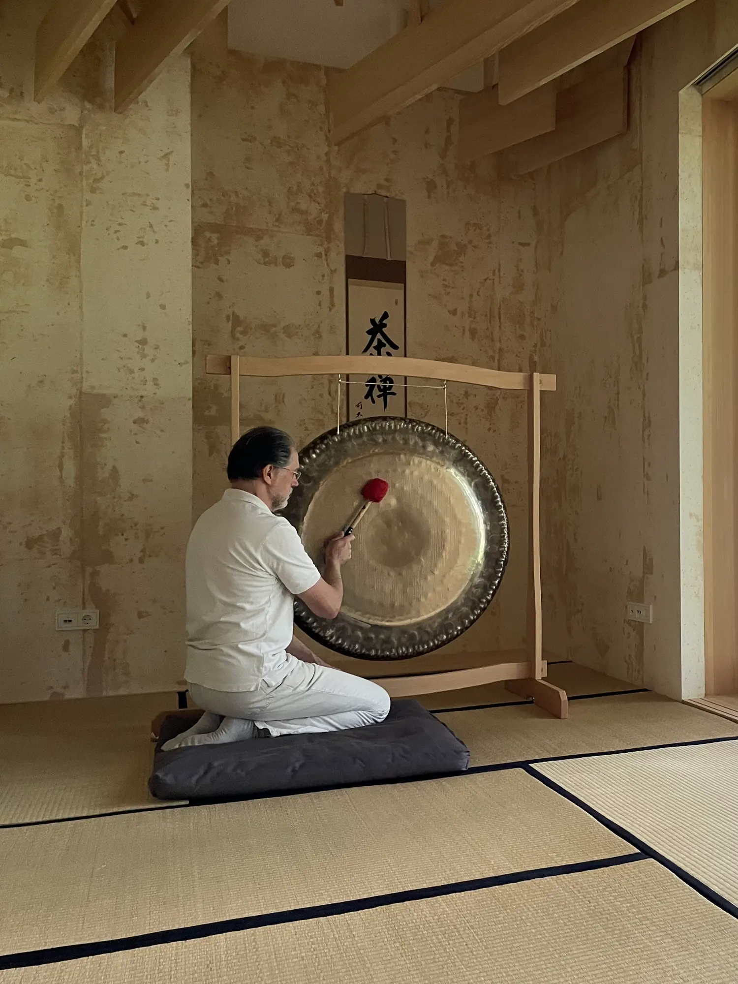 Alexander Renner kniet im Tatami-Raum seitlich am Gong, dahinter eine Kalligrafie-Rolle an der Lehmwand