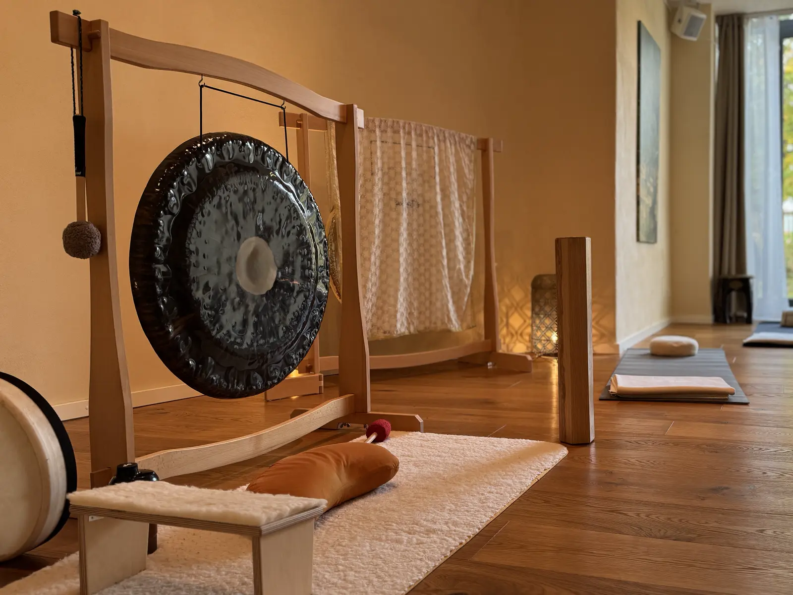 Dunkler Gong am hellen Holzständer in einem warm beleuchteten Raum, davor Meditationsbänkchen und Kissen, im Hintergrund ausgelegte Matten für die Gruppe