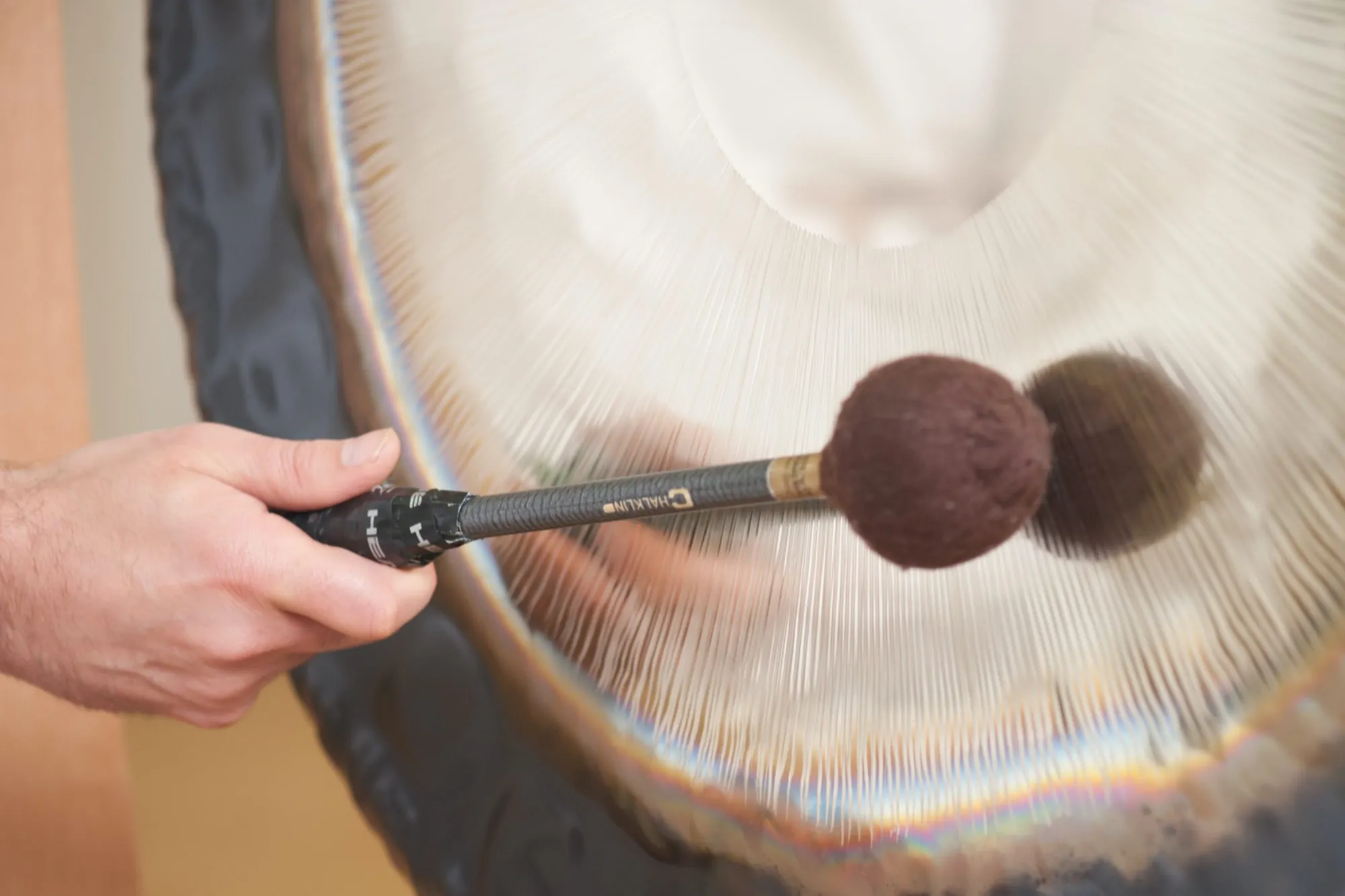 Hand führt einen Haklin-Schlägel kraftvoll über einen Gong mit Sonnenschliff, sichtbare Bewegung